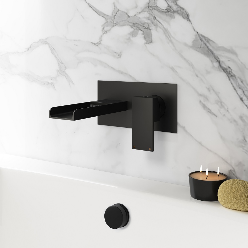A matt black, wall mounted waterfall bath filler tap against a marble-effect tiled wall, with a lit candle and loofah on the edge of a bathtub, exuding modern elegance.