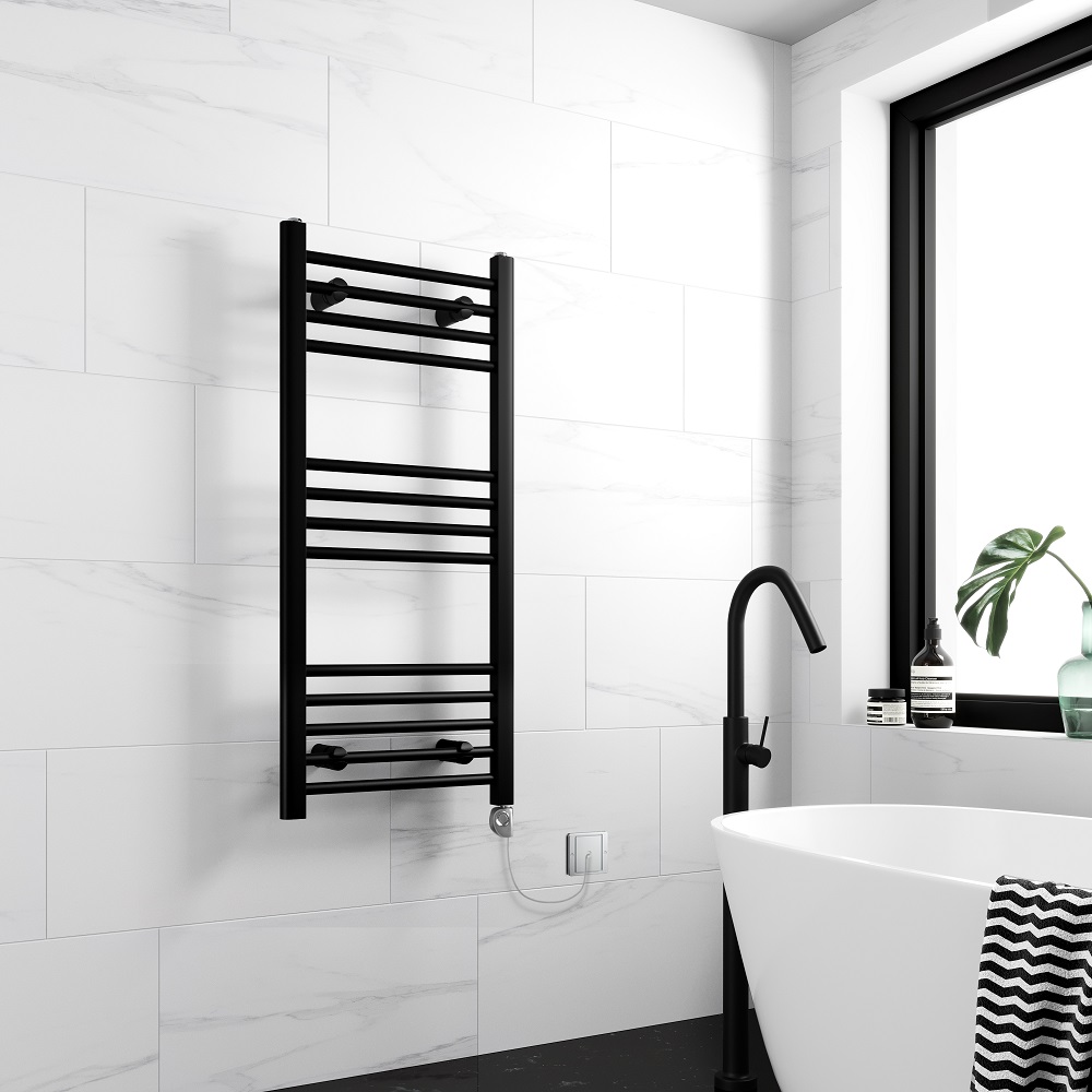 A matt black straight heated towel rail mounted on a marble effect tiled wall in a modern bathroom, with a freestanding bathtub and black faucet in view. 