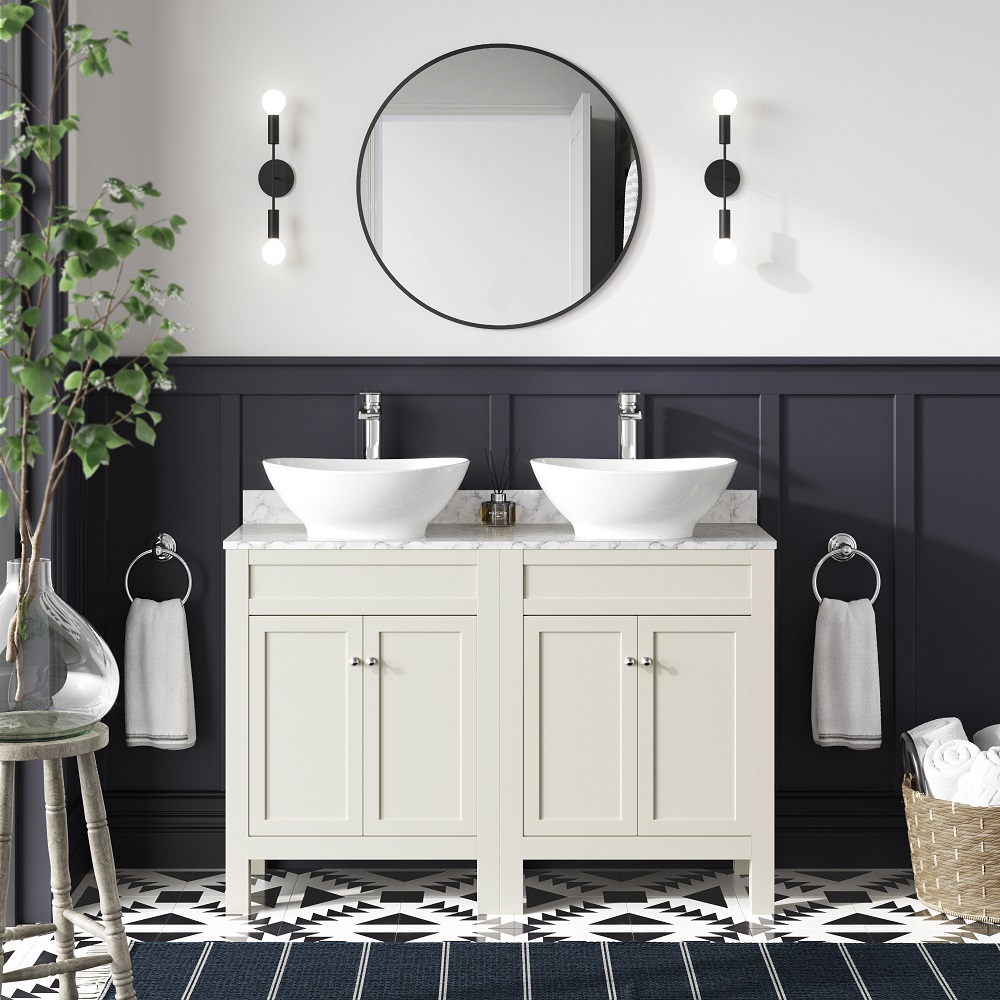 A stylish bathroom vanity area with two chalk white cabinets topped with marble counters and vessel basins, against a navy blue wainscoted wall. A round mirror flanked by modern wall sconces adds to the chic decor. The floor is adorned with bold black and white geometric tiles. ad accessories like towels and plants give the space a fresh and inviting feel. 