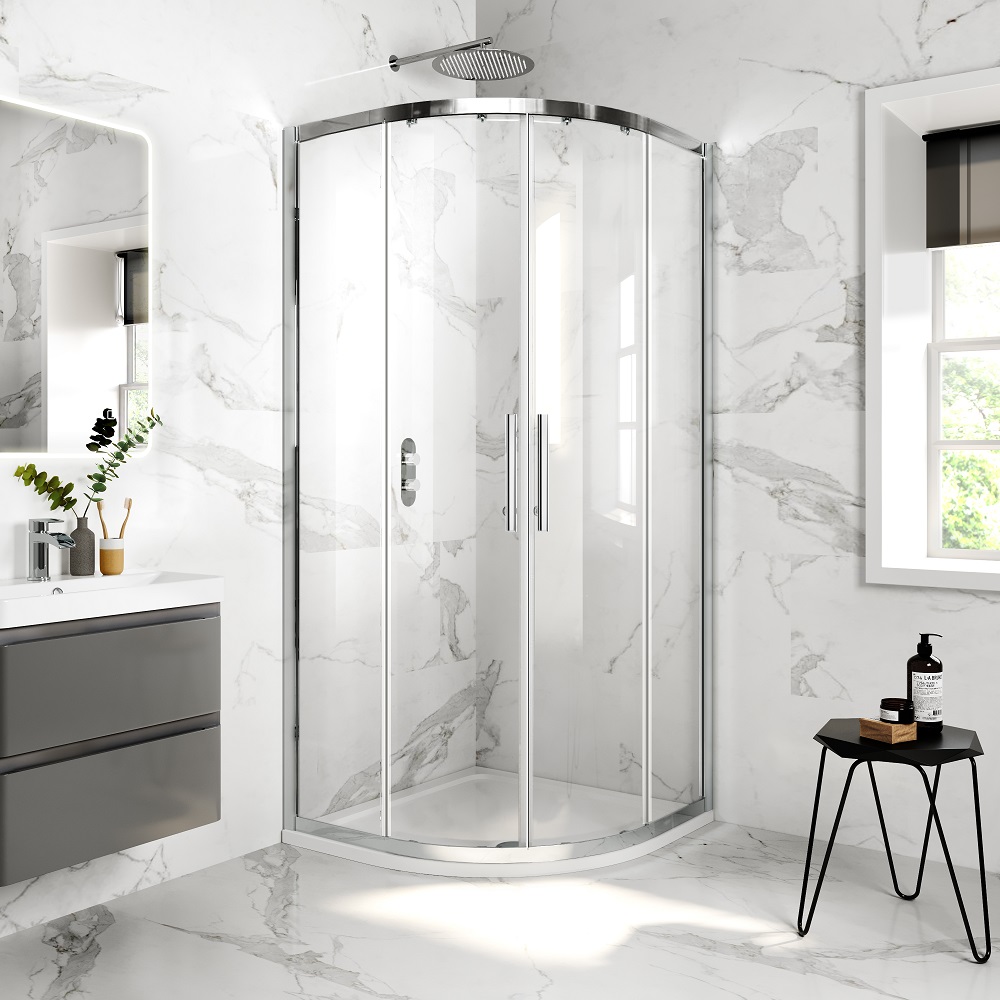 A modern corner shower with sliding doors set against white marble tiles. To the left is a grey vanity and to the right, a black side table with bath products. Natural light streams in from a window. 