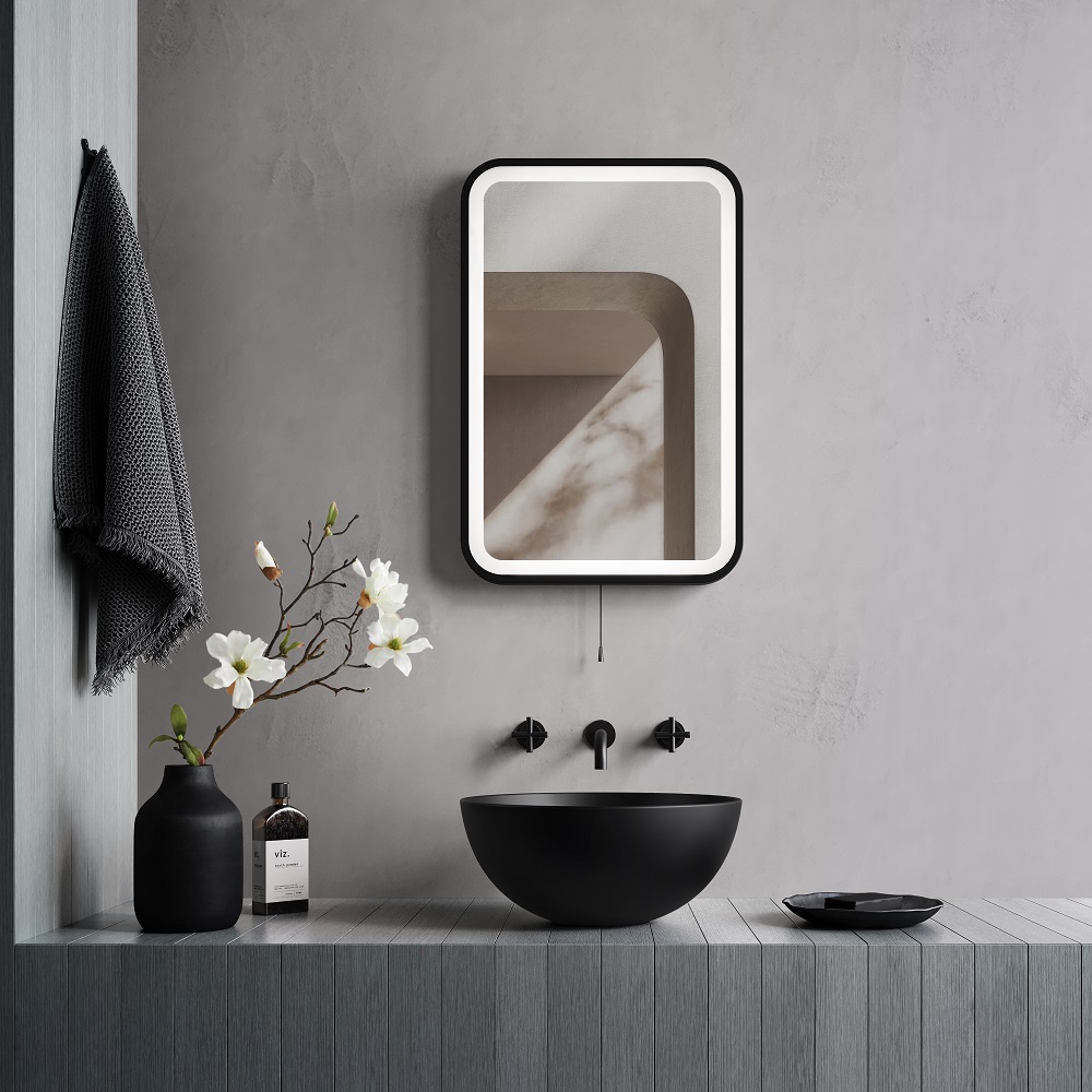 a chic bathroom vanity area with a matt black basin and wall-mounted faucets. Above it hangs a black framed illuminated LED mirror, reflecting a textured wall. To the side, a dark vase with white flowers and a grey towel add a touch of elegance.