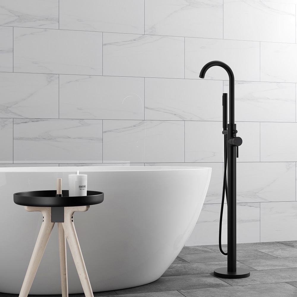 A modern bathroom featuring a sleek, freestanding bath tub with a matt finish, paired with a tall, matt black freestanding bath shower mixer tap. Walls are clad in large gloss tiles with a light marble effect, while floor is tiled with darker stone effect tiles. 