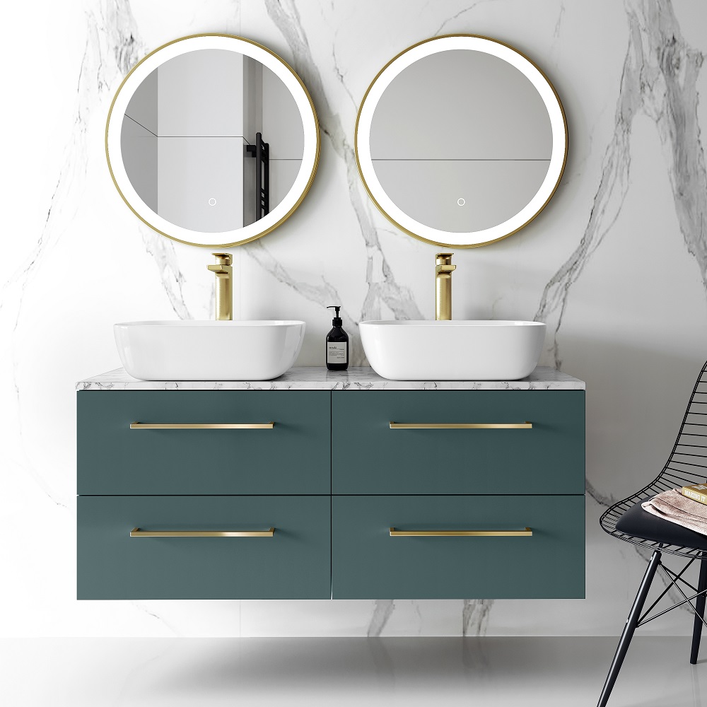 A modern bathroom vanity featuring a double wall-hung cabinet in midnight green with a marble countertop. Above the vanity are two round mirrors with gold accents, matching the gold faucets of the two white curved basins. A black soap dispenser sits on the countertop, and a black wire chair is partially visible to the side. 