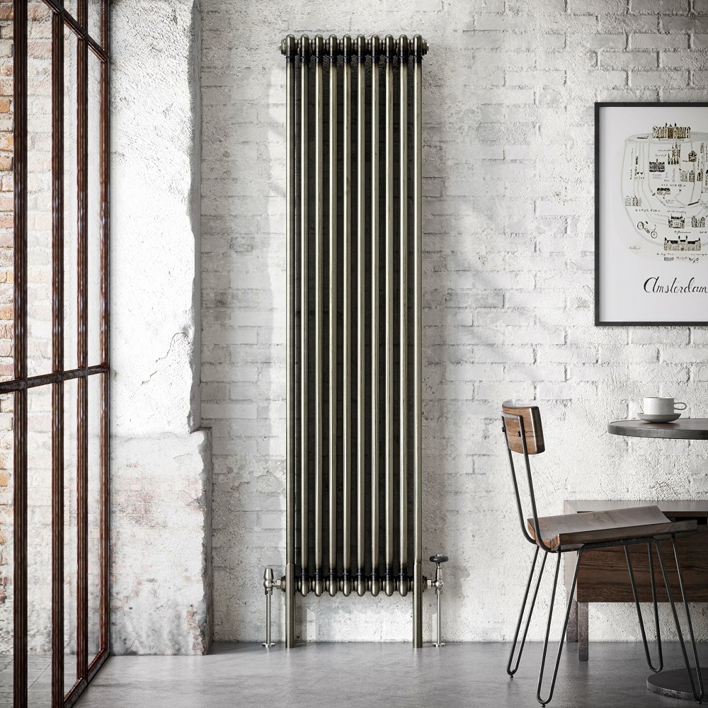 Chic and industrial inspired space with a tall raw metal triple column vertical radiator against a textured white brick wall. The room features urban loft elements such as large multi-pane window, a minimalistic wooden chair with black metal legs, and a simple wooden table set with a cup and saucer. A framed poster adds a touch of sophistication to the raw interior design. 