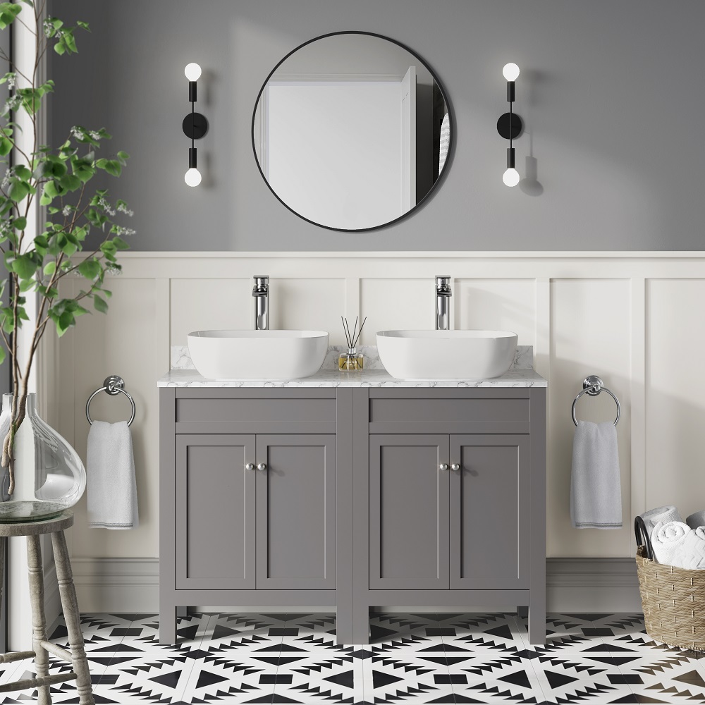 A grey double basin vanity unit with a white marble top and two countertop basins. A round mirror is centred above the vanity, flanked by two modern wall sconces. The room features wood panels and a bold black and white geometric floor tile. To the left, a glass vase with greenery adds a touch of nature. 