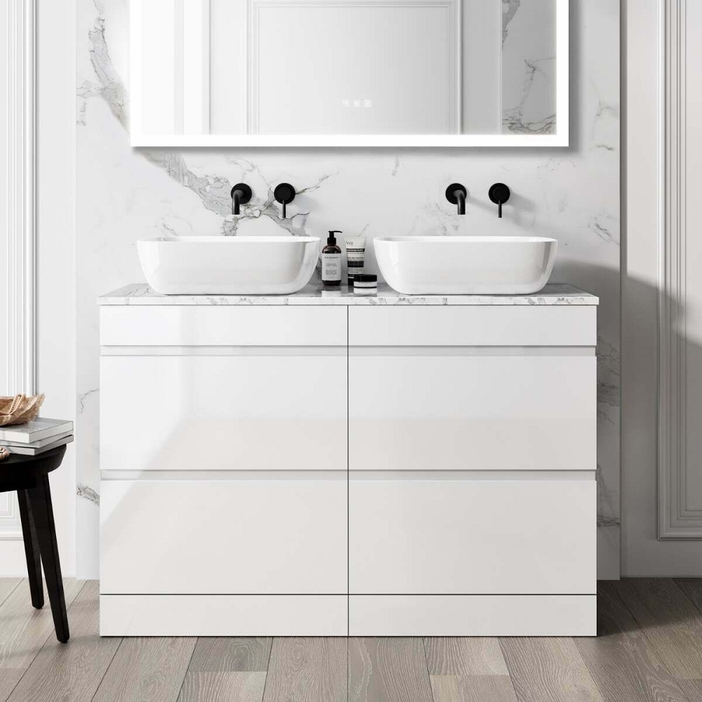 A modern gloss white double vanity with a marble top and two above-counter basins. The basins are paired with black faucets, and a black soap dispenser is placed between them. A large rectangular mirror with an illuminated frame is mounted above the vanity. The vanity stands against a marble wall, and the room has a light wooden floor. 