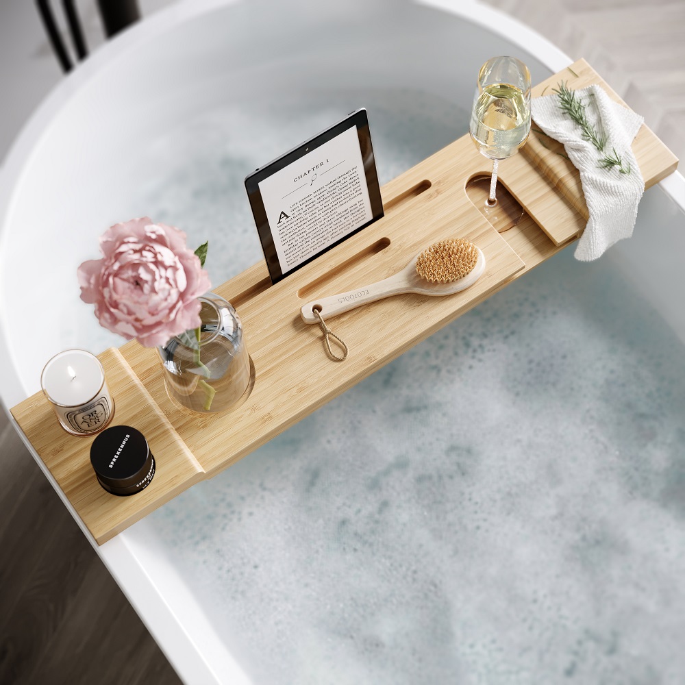 A bamboo bath caddy tray across a white freestanding tub filled with bubbles; includes a tablet displaying text, a glass of white wine, a pink peony, a clear glass with a single green leaf, a lit candle, a body brush, and a folded white towel.