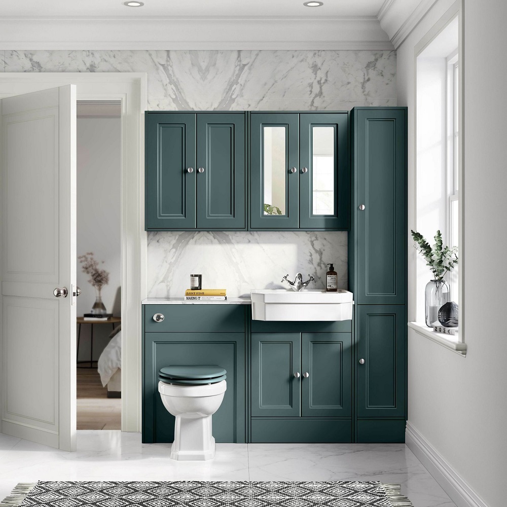 Timelessly styled bathroom featuring a deep green vanity cabinet with a luxurious marble countertop and white basin, accompanied by a classic white toilet. The design is complemented by marble wall tiles, a geometric patterned floor rug, and tasteful decor including a glass vase with greenery and artisanal soap bottles, all under natural light from a window. 