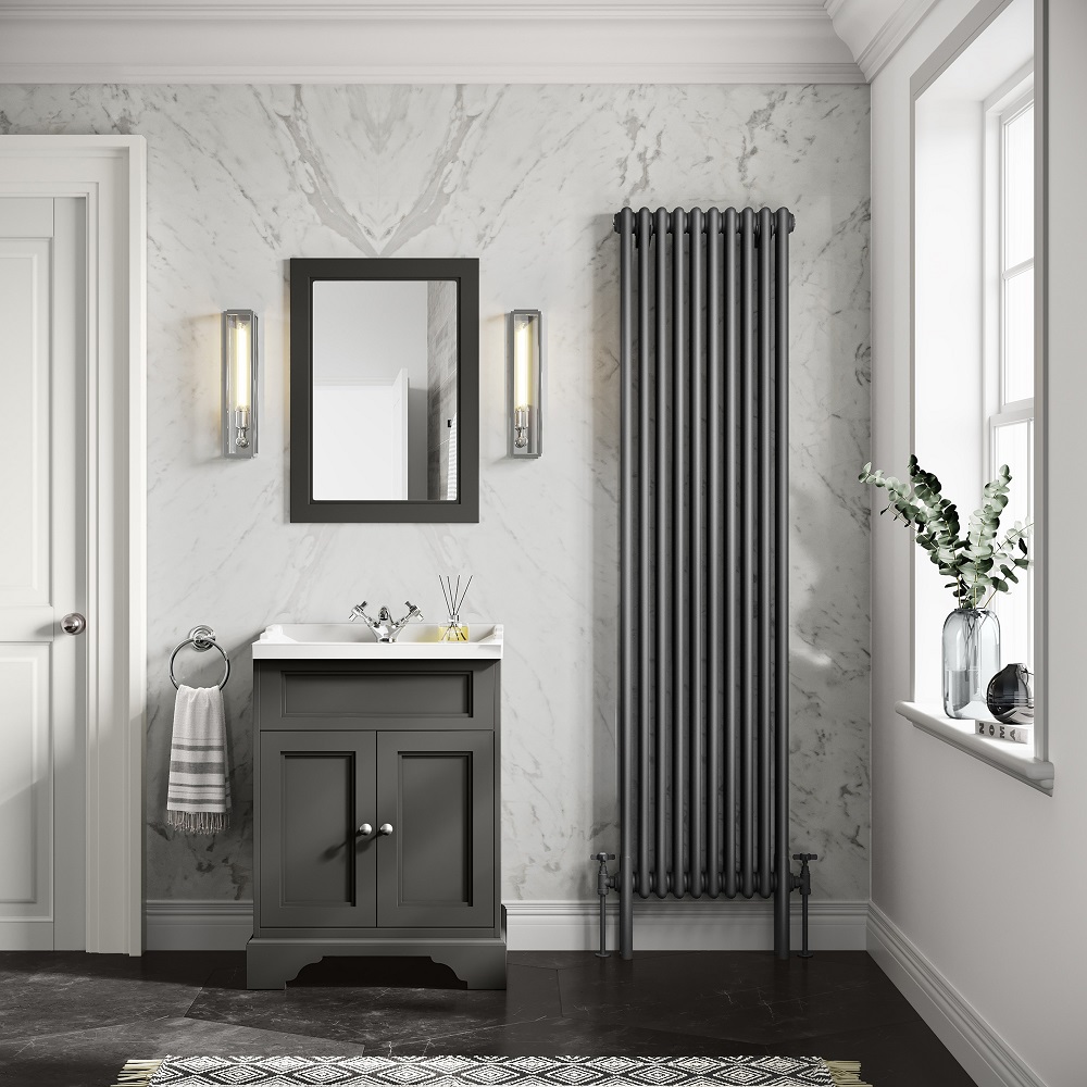 A classic bathroom design with a tall anthracite vertical radiator, marble wall, and a dark grey vanity cabinet topped with a white sink. Above the sink is a rectangular mirror flanked by wall-mounted lights. To the right, a window sill hosts a clear vase with greenery, and to the left, a striped hand towel hangs on a hook. The floor features a decorative black and white patterned rug. 