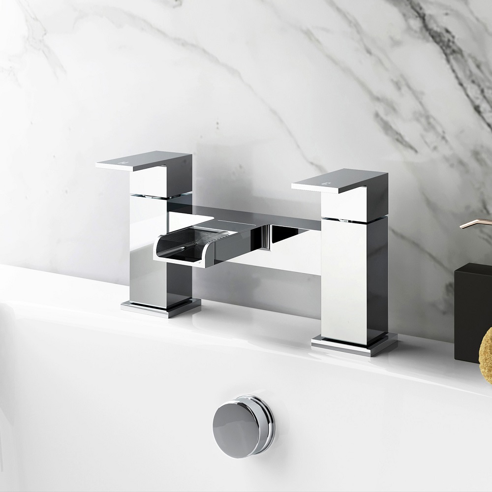 A modern chrome waterfall bath filler mounted on a white bathtub with a marble background, featuring geometric design, sleek lines, and a reflective surface that enhances the luxurious feel of the bathroom. 