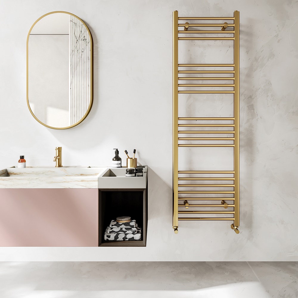 A contemporary bathroom with a brushed brass straight heated towel rail on a white marble wall, an oval brass-framed mirror above a pink and marble sink vanity with brass fixtures, and assorted toiletries, including a soap dispenser and brushes. A black storage cube under the vanity holds neatly folded towels with a patterned design. 