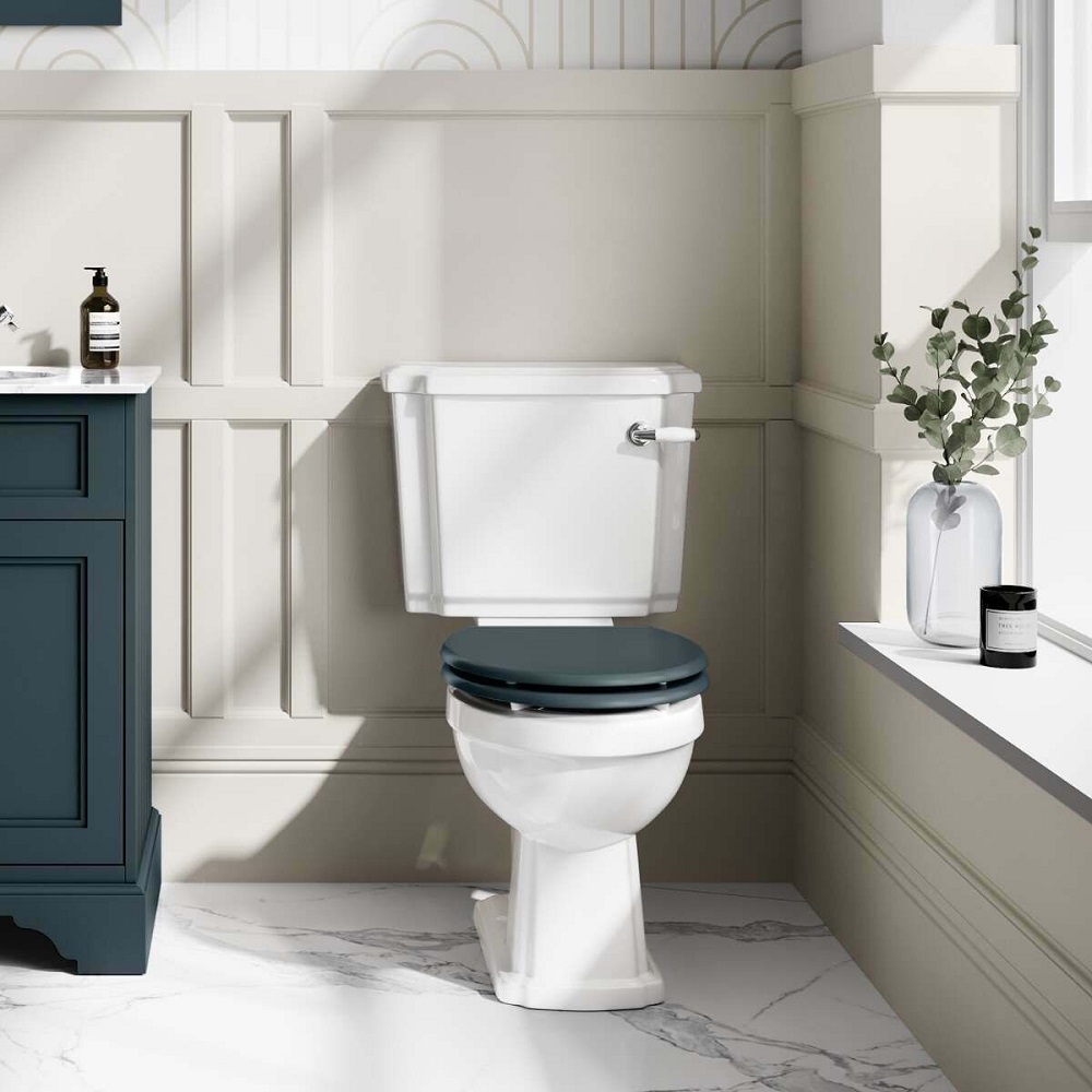 A bright and airy traditional bathroom featuring a close-coupled white toilet with a sophisticated blue wooden seat. The toilet is set against a backdrop of elegant cream wainscoting and marble flooring, with minimalist decor including a small clear glass vase with eucalyptus branches and a black jar, adding a touch of modernity to the classic space.