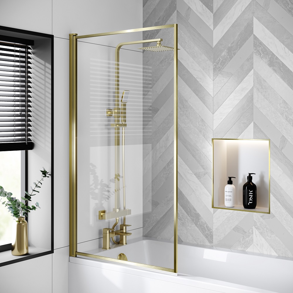 A brushed brass-framed bath screen with a hinged door, set agaist a herringbone-patterned marble wall. The screen is part of a shower over a bathtub, complete with a brass rainfall shower head, matching faucet, and controls. A recessed shelf holds luxury black and white labelled bath products, complemented by a potted plant and a vase on a ledge. 