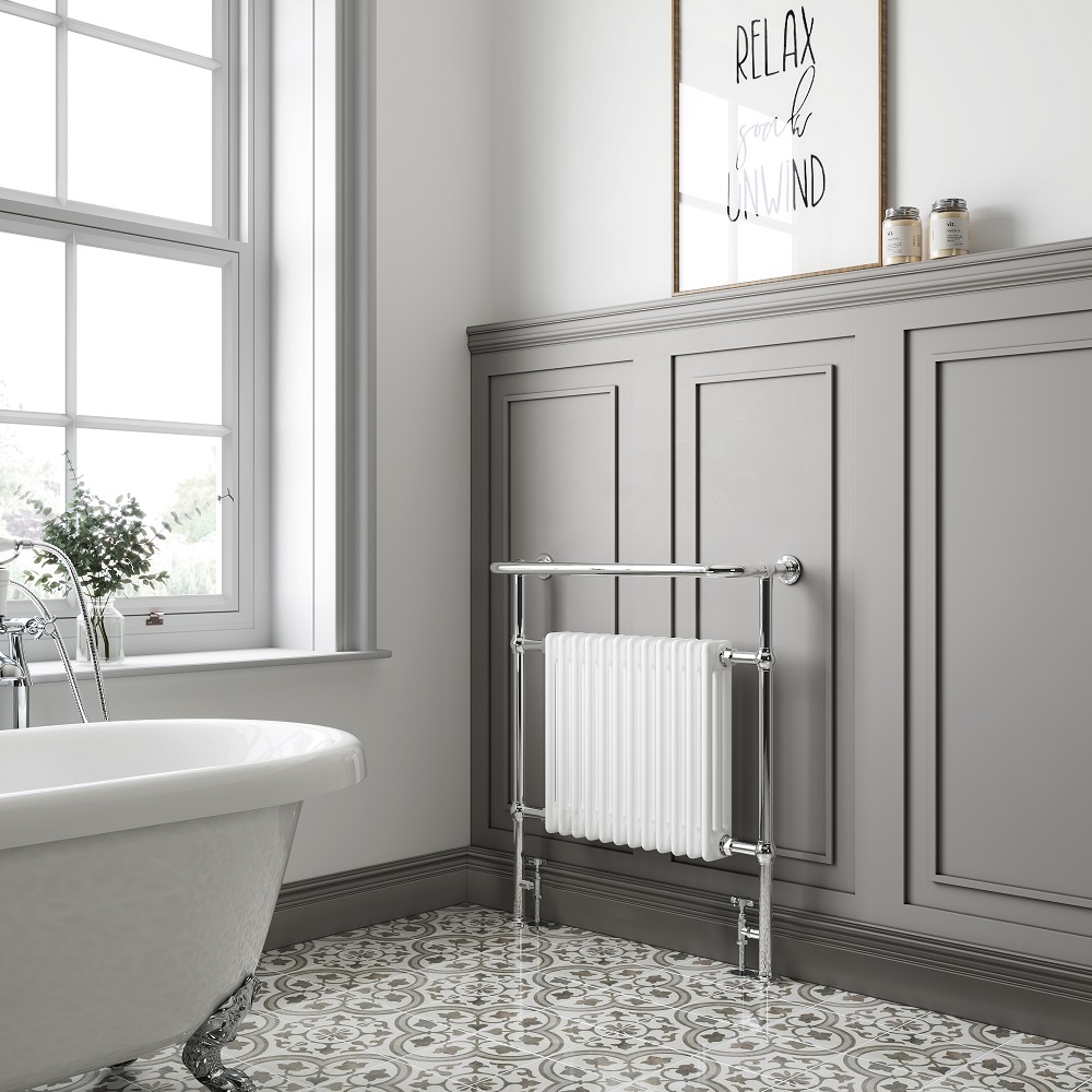 Traditional white heated towel radiator with a chrome overhanging rail, positioned against a grey panelled wall in a light-filled bathroom with ornate patterned floor tiles. A framed calligraphy quote 'Relax, Soak, Unwind' invited a peaceful atmosphere, enhancing the room's classic comfort and style. 
