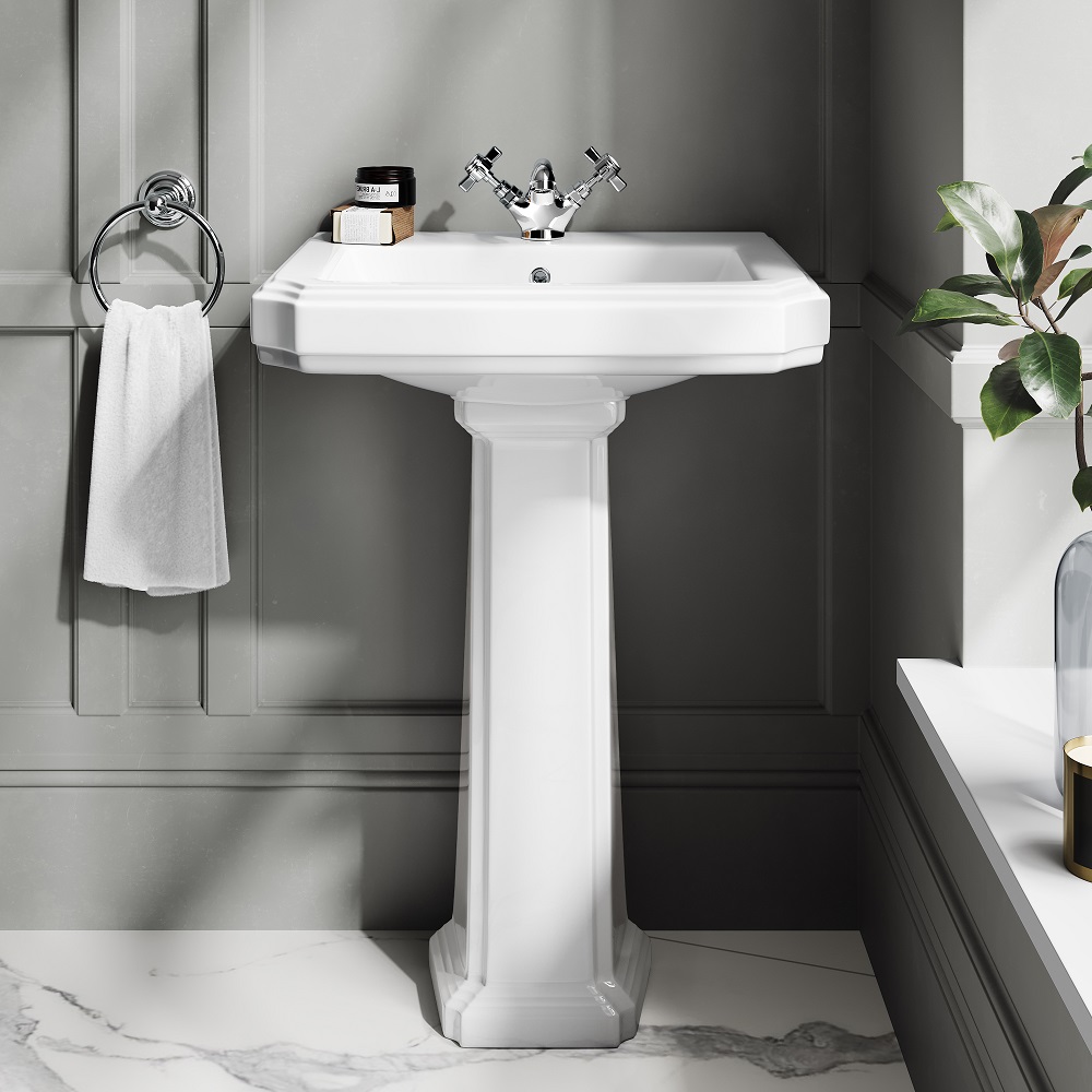 Timeless white pedestal basin with classic chrome taps, set against a grey panelled all for a sophisticated contrast. The design is complemented by a silver towel ring and marble floor, with a touch of greenery adding freshness to the elegant bathroom space.