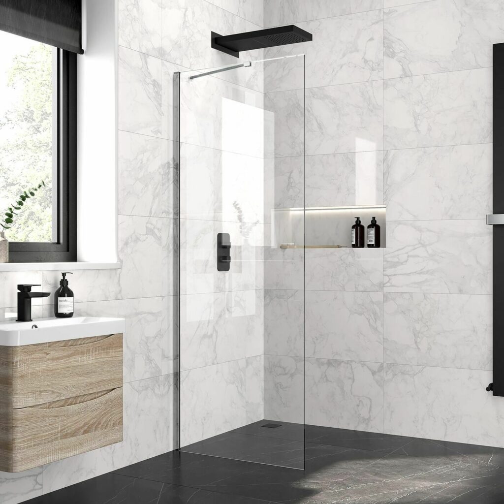 Modern marble tiled bathroom featring a sleek wet room shower glass panel with a matt black overhead shower system, adjacent to a wooden vanity with black fixtures and natural light from a large window. 