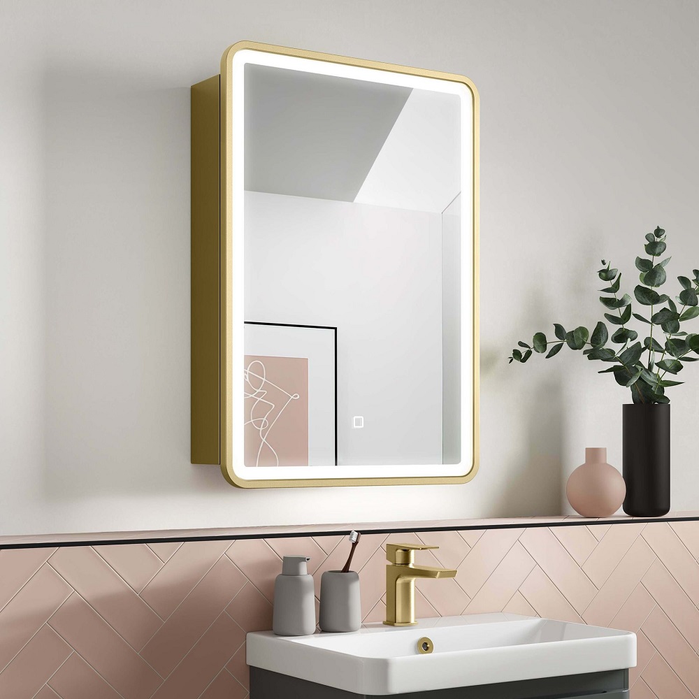 Elegant bathroom interior featuring a brushed brass LED mirror cabinet above a white sink with matching brass tap, set against blush pink herringbone tiles and minimalist decor.