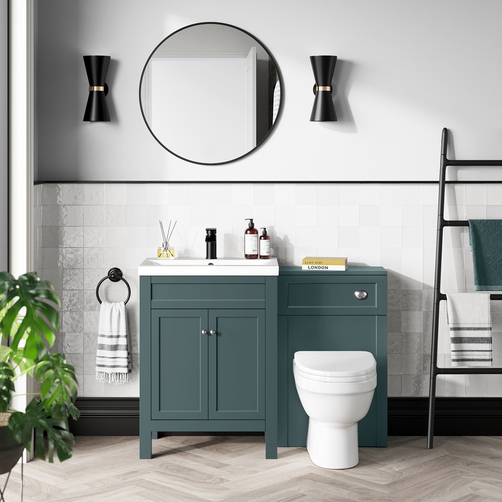 A serene bathroom setup featuring a midnight green vanity with a white countertop basin, complemented by a round mirror and dual black wall sconces. The space is accented with a sleek black towel ladder, a white toilet, and decorative elements like a reed diffuser, hand soap and neatly stacked books, creating a cosy and inviting atmosphere. A potted plant adds a touch of greenery, enhancing the rooms fresh and clean feel. 