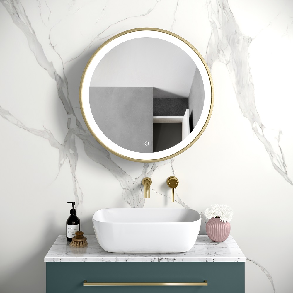 A luxurious bathroom vignette showcasing a sleek rectangular vessel sink atop a marble countertop with a matt teal vanity unit. Above, a round illuminated LED Mirror framed in matt brass casts a soft glow, enhancing the marble's elegant veining. This scene is accessorised with a black soap dispenser, a brass brush and a delicate pink vase with white pompom flowers, evoking a sense of sophisticated and modern ambiance. 