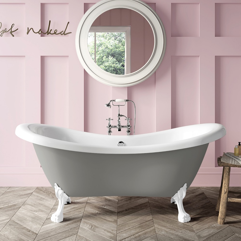 Romantic and whimsical bathroom featuring a dove grey roll-top bath with contrasting white ornate feet, set against a soft pink panelled wall with playful 'get naked' script, a classic round mirror, and warm herringbone wooden floor, inviting a touch of elegance and fun to the bathing experience. 