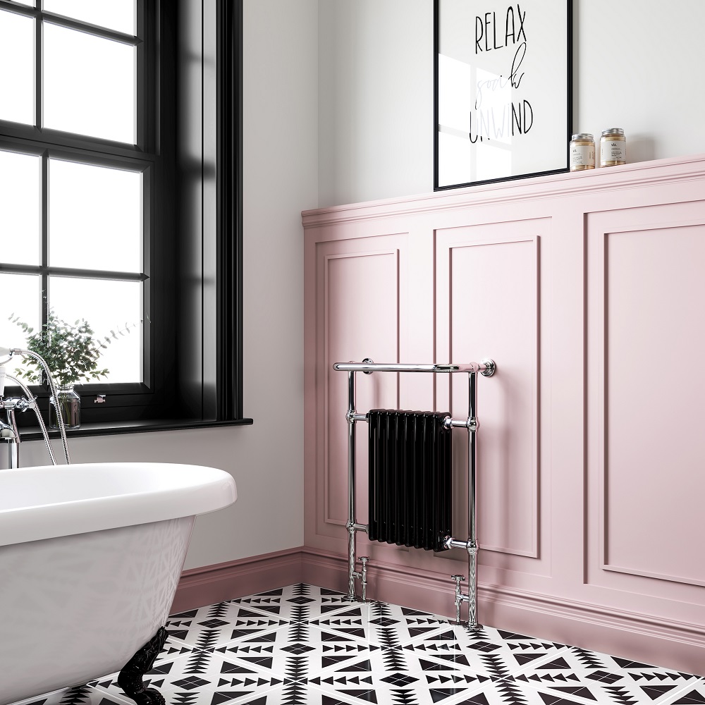 Luxurious white freestanding bathtub in a room with chic pink panelling and bold monochrome geometric floor tiles, featuring a classic black framed 'Relax, soak, unwind' quote, natural light from large windows, and an elegant black traditional column towel radiator.