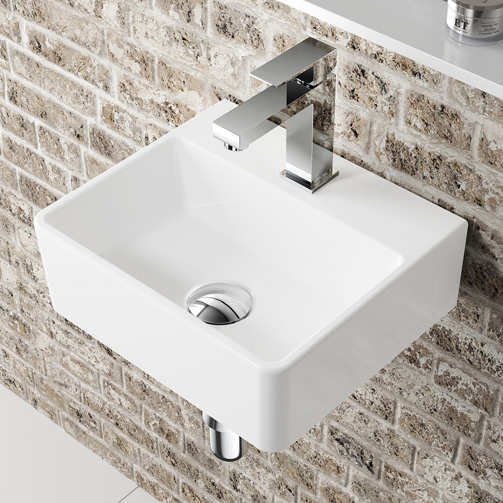 A minimalist square wall-mounted cloakroom basin in white, paired with a sleek chrome mixer tap. The basins clean lines and angular form contrast beautifully with the brick wall behind it, blending contemporary design with industrial textures. The subtle shine of the tap and the basins smooth surface 