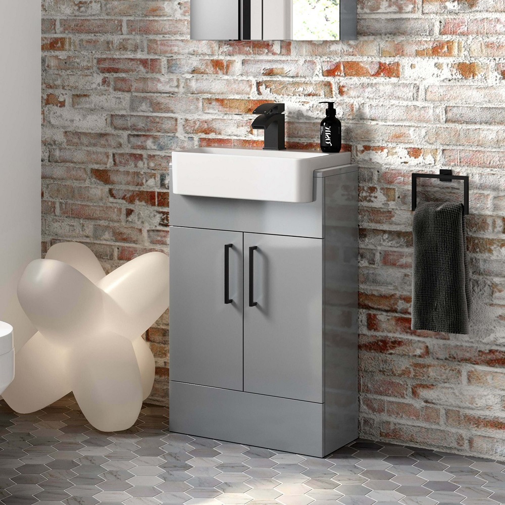 A modern grey vanity unit with a semi-recessed basin set against a rustic exposed brick wall. A matt black faucet complements the clean lines of the vanity, while a black soap dispenser adds a touch of contemporary elegance. The ensemble is situated on a geometric patterned floor, with a charcoal towel hanging neatly on a black towel holder, contributing to the urban-chic aesthetic of the space. A playful, abstract light sculpture on the side softens the industrial vibe with a hint of whimsy. 