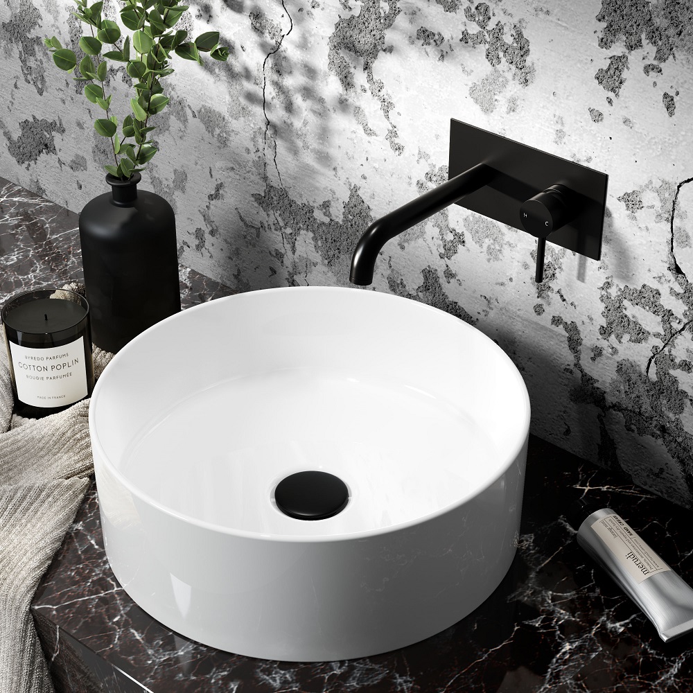 A stylish round countertop basin in pristine white, featuring a sleek black overflow ring, paired with a matt black wall-mounted faucet. The basin sits atop a marbled countertop with grey and black veining, complemented by a black vase holding green foliage, a plush beige towel, and a luxury hand soap, creating a chic and modern bathroom aesthetic. 