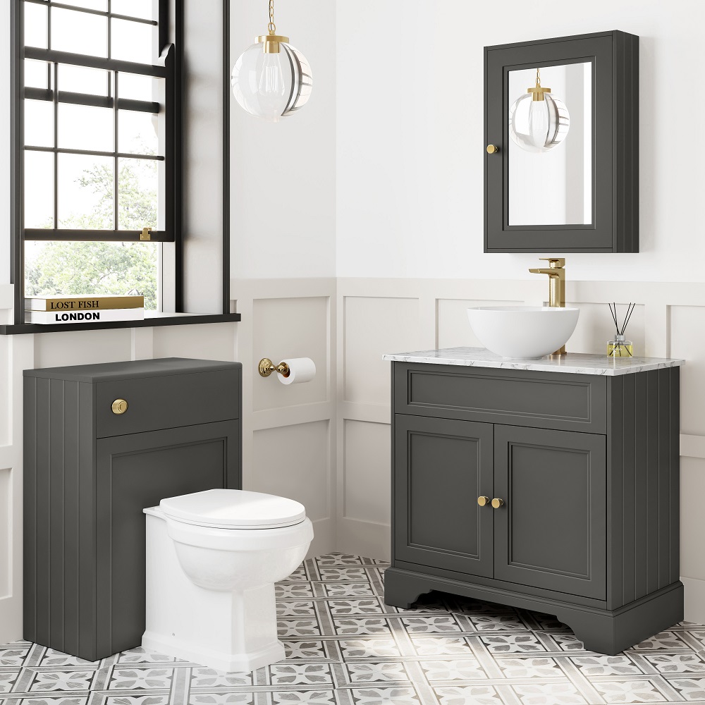 An elegant bathroom features a graphite grey vanity with a marble top and a round countertop basin, measuring 840mm, accented with gold fixtures. Beside it is a matching grey WC unit, creating a cohesive look. The room is brightened by natural light streaming through a large window and a chic pendant light. The patterned floor tiles add a touch of classic charm, making this setting an aspirational choice for those seeking the best sink for their bathroom.