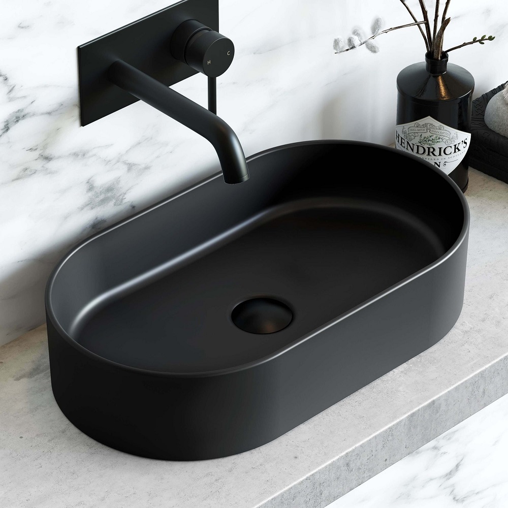 A Dakota matt black oval countertop basin measuring 525mm, presenting a bold statement atop a marble look surface. Its deep hue contrasts sharply with the grey and white marbled backdrop, complemented by a sleek black wall mounted faucet. A decorative vase with dried botanicals adds a touch of organic texture, enhancing the modern and sophisticated design of the bathroom setup. 