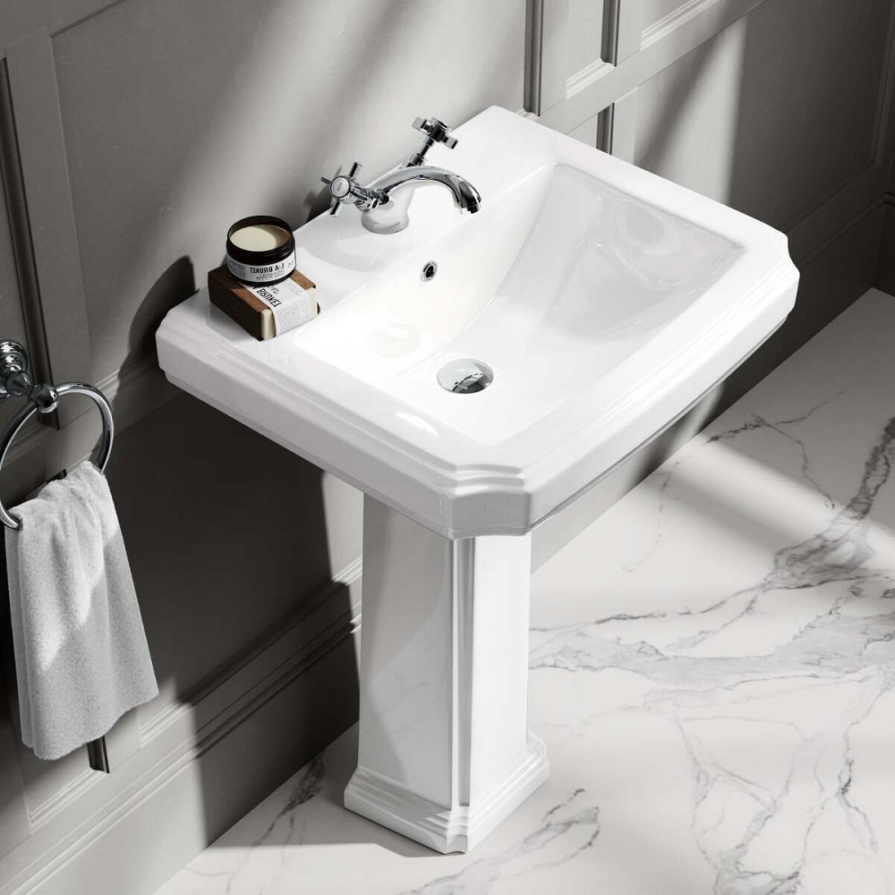 A traditional white pedestal basin with a single tap hole, showcased in a bathroom with classic charm. The basin features a spacious washing area and a graceful profile, standing against a wall with subtle grey tones that complement the white marble effect floor. A timeless chrome faucet, a jar of grooming essentials, and a neatly hung towel complete this elegant and functional setup. 
